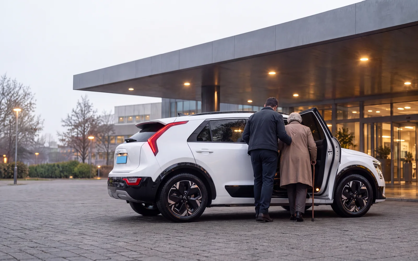 Illustratie: witte elektrische SUV bij de ingang van een ziekenhuis overdag, de chauffeur helpt een oudere passagier met stok, beiden van achteren